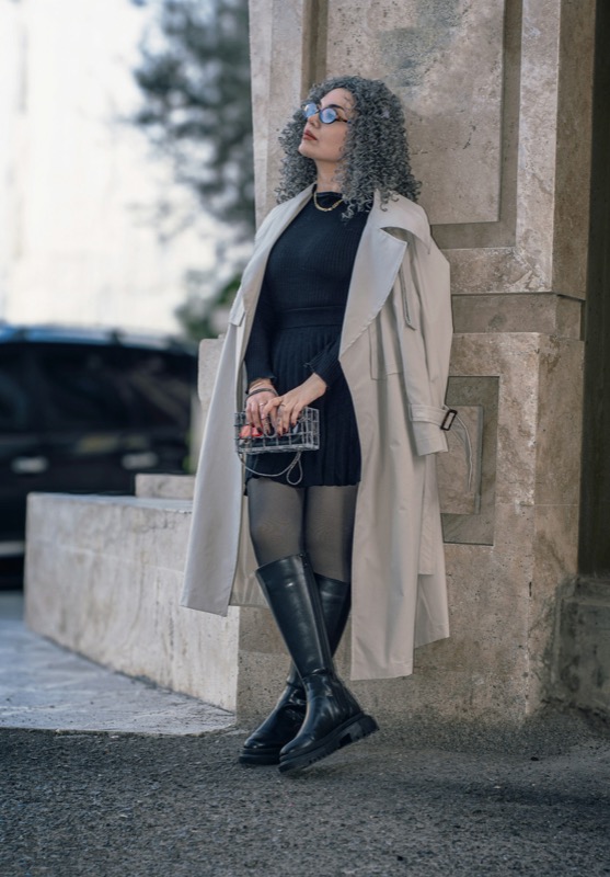 Kvinna i beige trenchcoat över svart klänning, mot en stenpelare